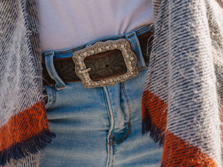 A woman with long dark hair has a brown belt with a large buckle and her hands are clasped behind her back.