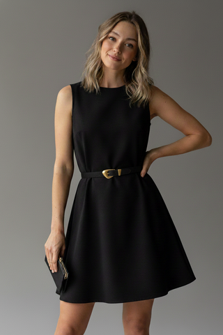 A woman wearing a white sleeveless top, blue jeans, and a brown belt with long blonde hair stands , smiling softly.