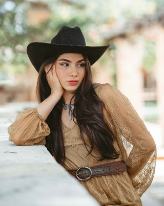 A woman with long dark hair has a brown belt with a large buckle and her hands are clasped behind her back.