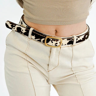 A woman with long hair wearing a black sleeveless top and blue jeans stands against a dark wall.